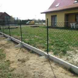 Muka Biuro Zarządzania Nieruchomościami - Siatka ogrodzeniowa z zielonymi słupkami i betonową podmurówką na piaszczystym terenie, dom w tle. Widoczny fragment posesji i osoba przy ogrodzeniu.