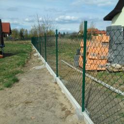 Muka Biuro Zarządzania Nieruchomościami - Fragment nowo postawionego ogrodzenia z siatki z zielonymi słupkami, widoczne bloczki betonowe i cegły na trawie. Fundament ogrodzenia betonowy.