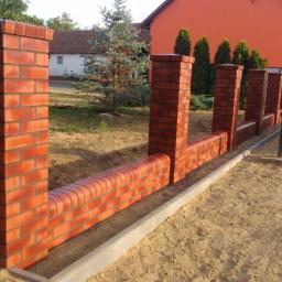 Muka Biuro Zarządzania Nieruchomościami - Ceglany murek ogrodzeniowy z filarami, w trakcie budowy, z widocznymi elementami konstrukcyjnymi i przygotowanym terenem, na tle domu i zieleni.