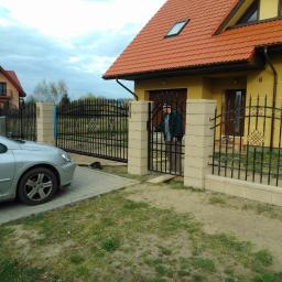 Muka Biuro Zarządzania Nieruchomościami - Nowoczesne ogrodzenie kute z bramą i furtką, z jasnymi słupkami z bloczków. Na podjeździe stoi srebrny samochód, w tle dom z czerwoną dachówką.