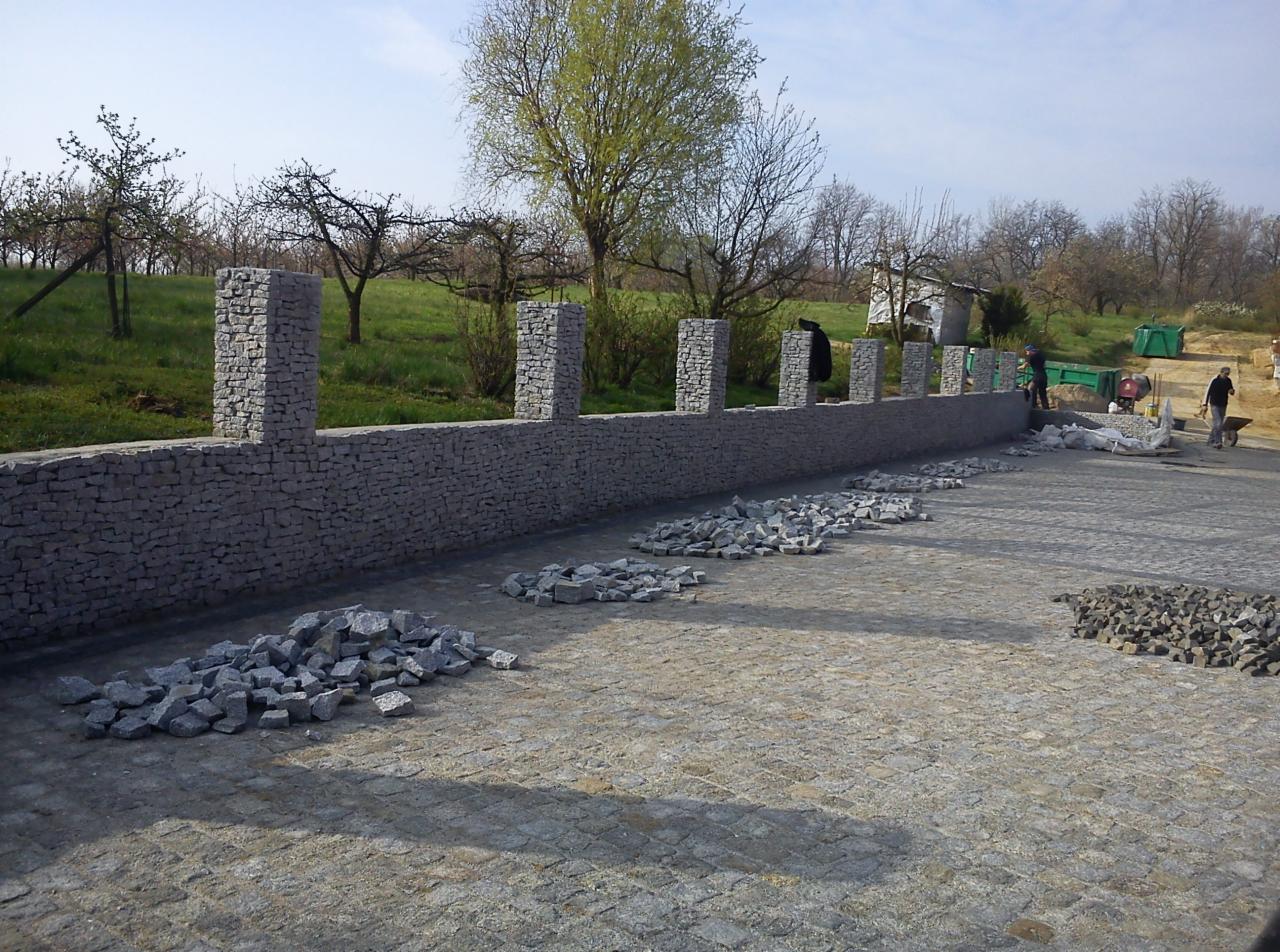 Budowa ogrodzenia z kamienia gabionowego z filarami, widoczne narzędzia i materiały budowlane, w tle zieleń i drzewa.