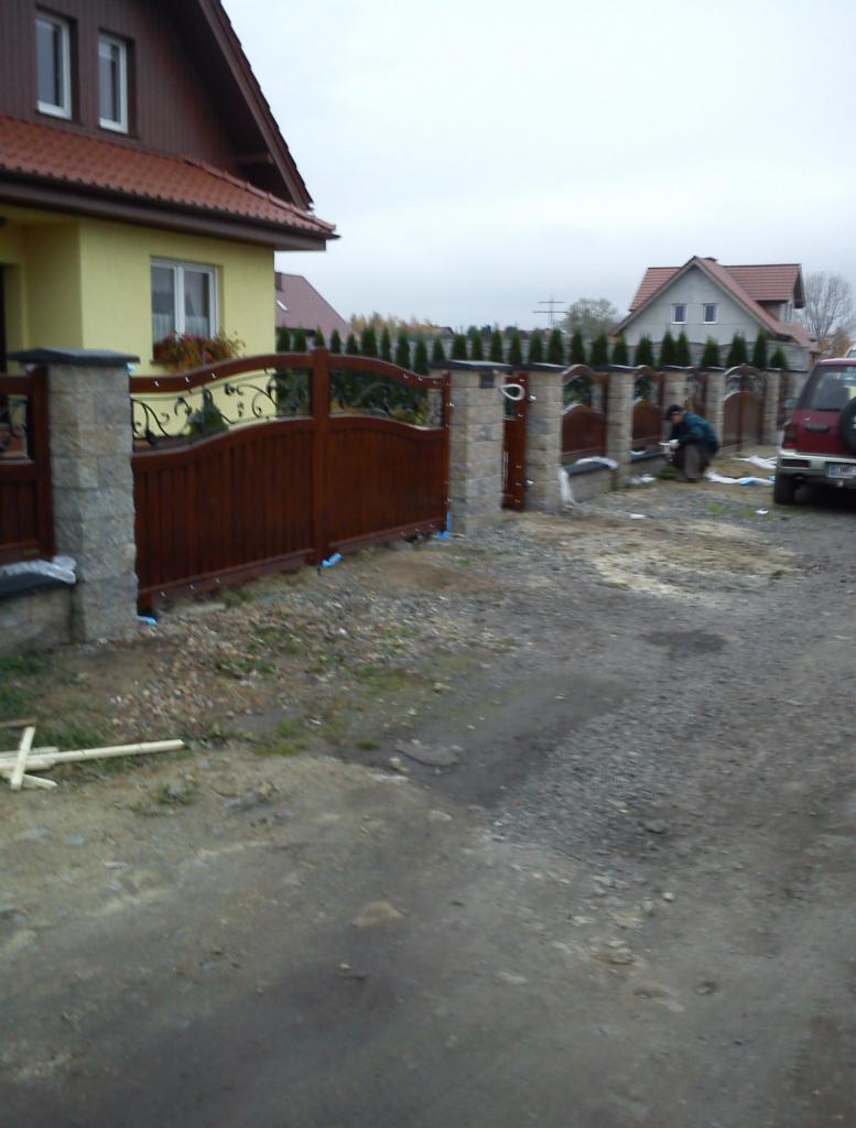 Ciemnobrązowe, drewniane ogrodzenie z kamiennymi słupkami. Na drugim planie widać osobę pracującą przy ogrodzeniu, a w tle domy i drzewa.