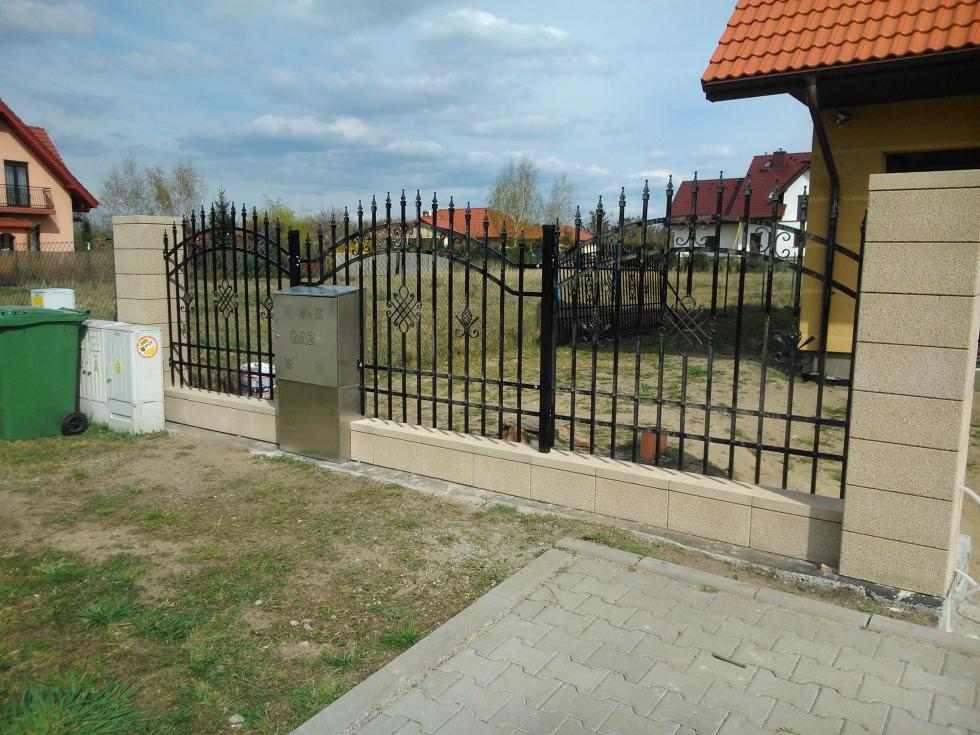 Kute, czarne ogrodzenie z beżowymi słupkami z bloczków betonowych. Na słupku skrzynka na gaz z numerem domu. W tle domy z czerwonymi dachami i błękitne niebo.