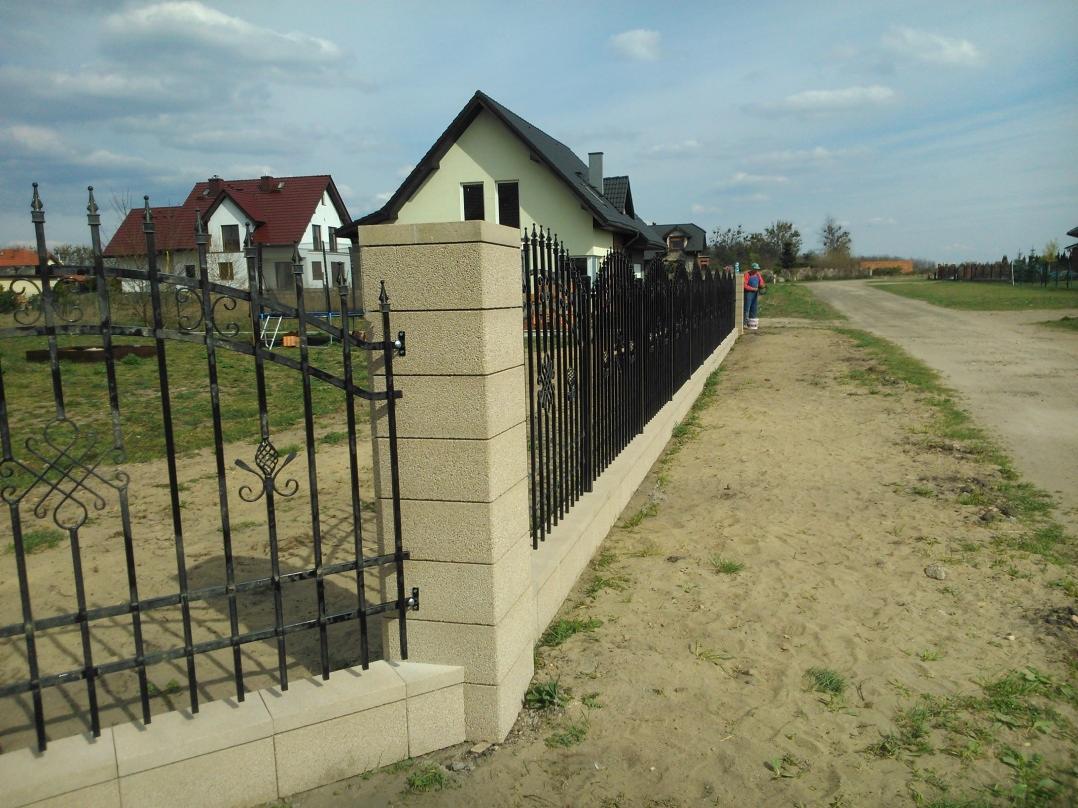 Eleganckie ogrodzenie kute z elementami dekoracyjnymi, osadzone na słupkach z jasnego kamienia, w tle domy jednorodzinne i polna droga.