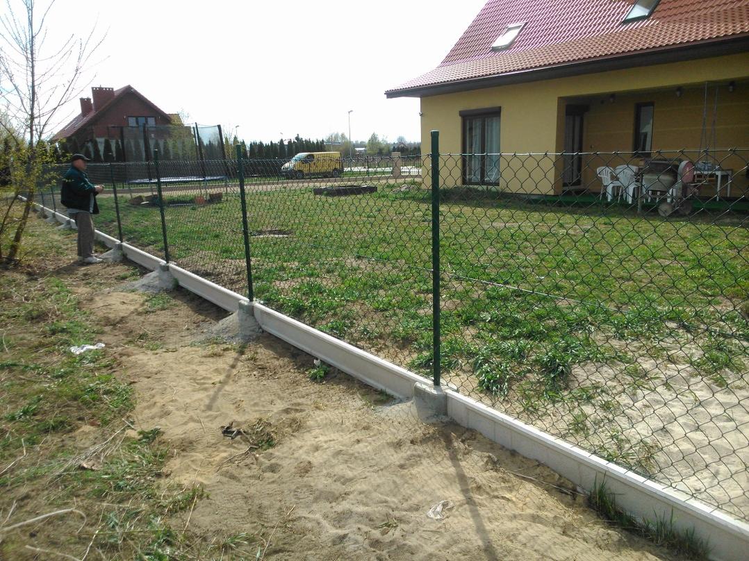 Siatka ogrodzeniowa z zielonymi słupkami i betonową podmurówką na piaszczystym terenie, dom w tle. Widoczny fragment posesji i osoba przy ogrodzeniu.