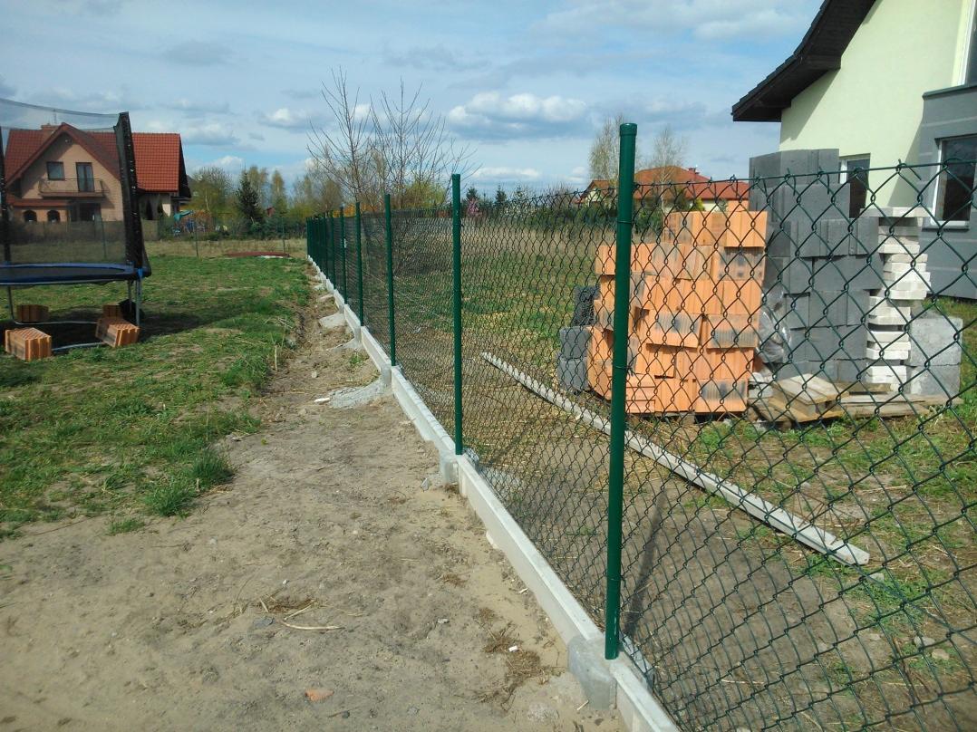 Fragment nowo postawionego ogrodzenia z siatki z zielonymi słupkami, widoczne bloczki betonowe i cegły na trawie. Fundament ogrodzenia betonowy.