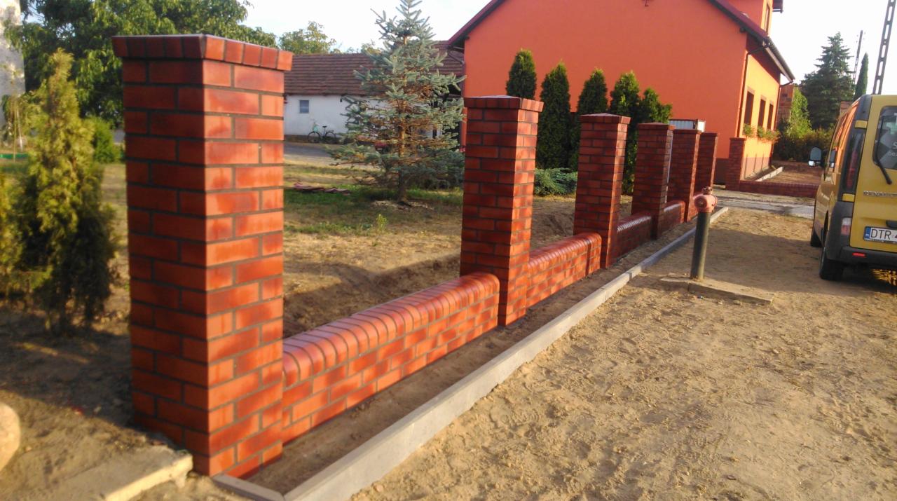 Ceglany murek ogrodzeniowy z filarami, w trakcie budowy, z widocznymi elementami konstrukcyjnymi i przygotowanym terenem, na tle domu i zieleni.
