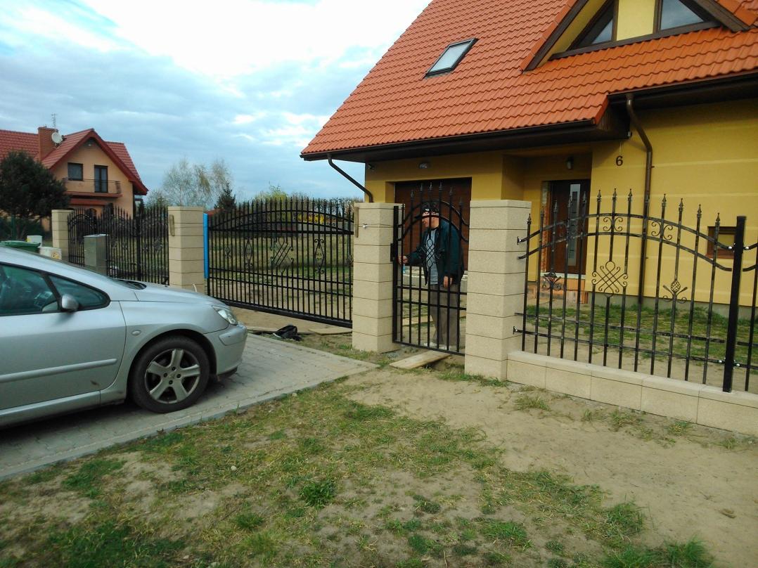 Nowoczesne ogrodzenie kute z bramą i furtką, z jasnymi słupkami z bloczków. Na podjeździe stoi srebrny samochód, w tle dom z czerwoną dachówką.
