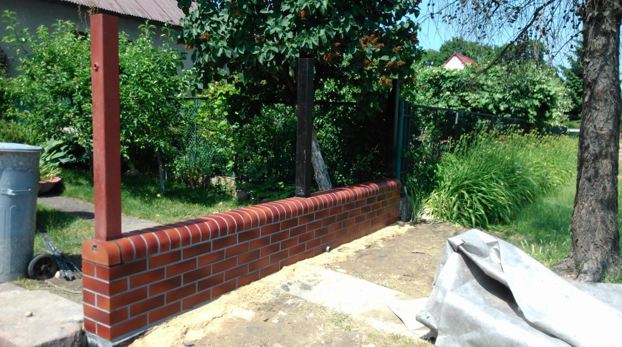 Ceglany murek ogrodzeniowy z filarami, w tle zieleń ogrodu. Widoczne narzędzia i materiały budowlane.