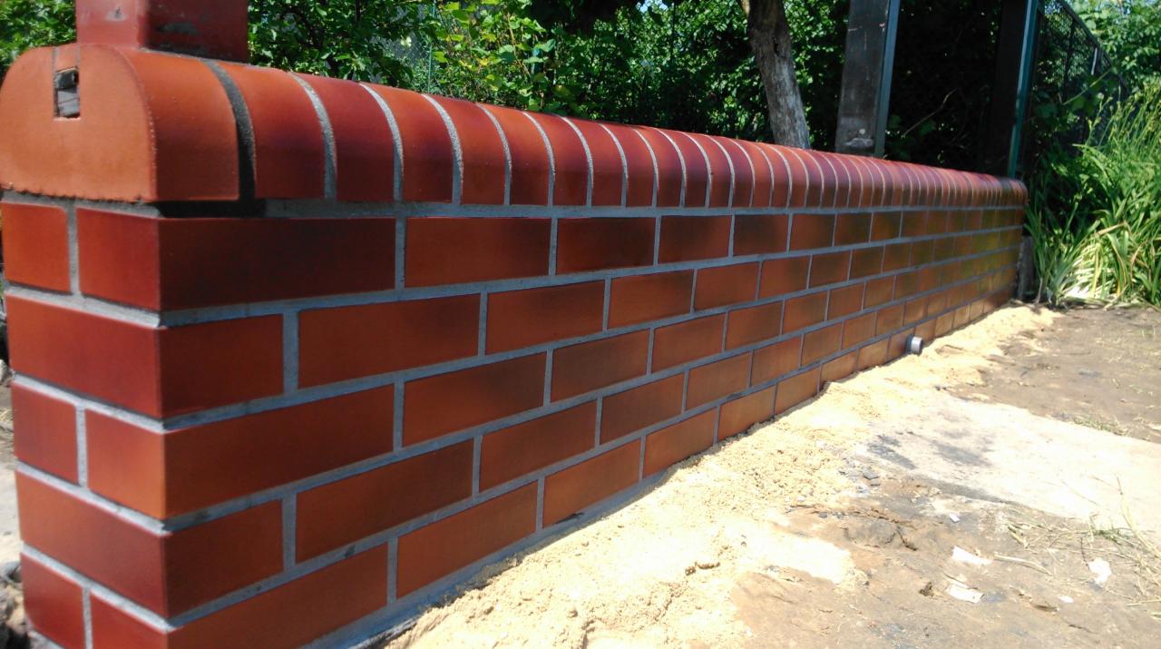 Mur z czerwonej cegły klinkierowej, wykończony półokrągłymi elementami na górze. Widoczne spoiny i fragment zieleni w tle. Nietypowe wykończenie muru.
