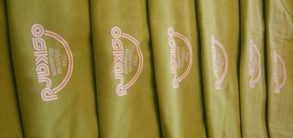 Rząd oliwkowych koszulek z identycznym, jasnoróżowym nadrukiem 'Tyska Spółdzielnia Mieszkaniowa' w pionowym układzie na każdej z nich.