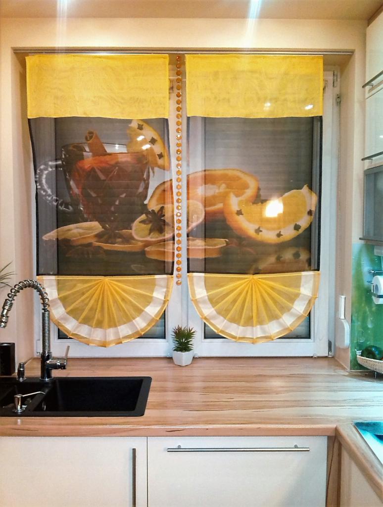 Żółto-czarne zasłony panelowe z motywem owocowym w kuchni, zawieszone na oknie z dekoracyjnym sznurem koralików. Widoczny zlew i blat.
