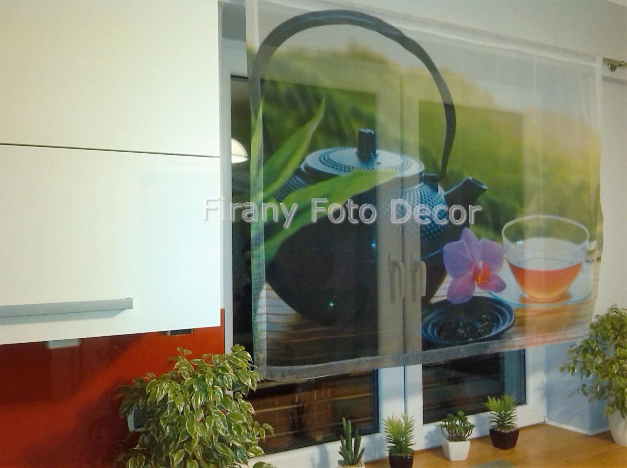 Zasłona roleta z nadrukiem czajnika i filiżanki herbaty, zamontowana w oknie kuchni. Widoczne rośliny doniczkowe i fragment szafek kuchennych w tle.