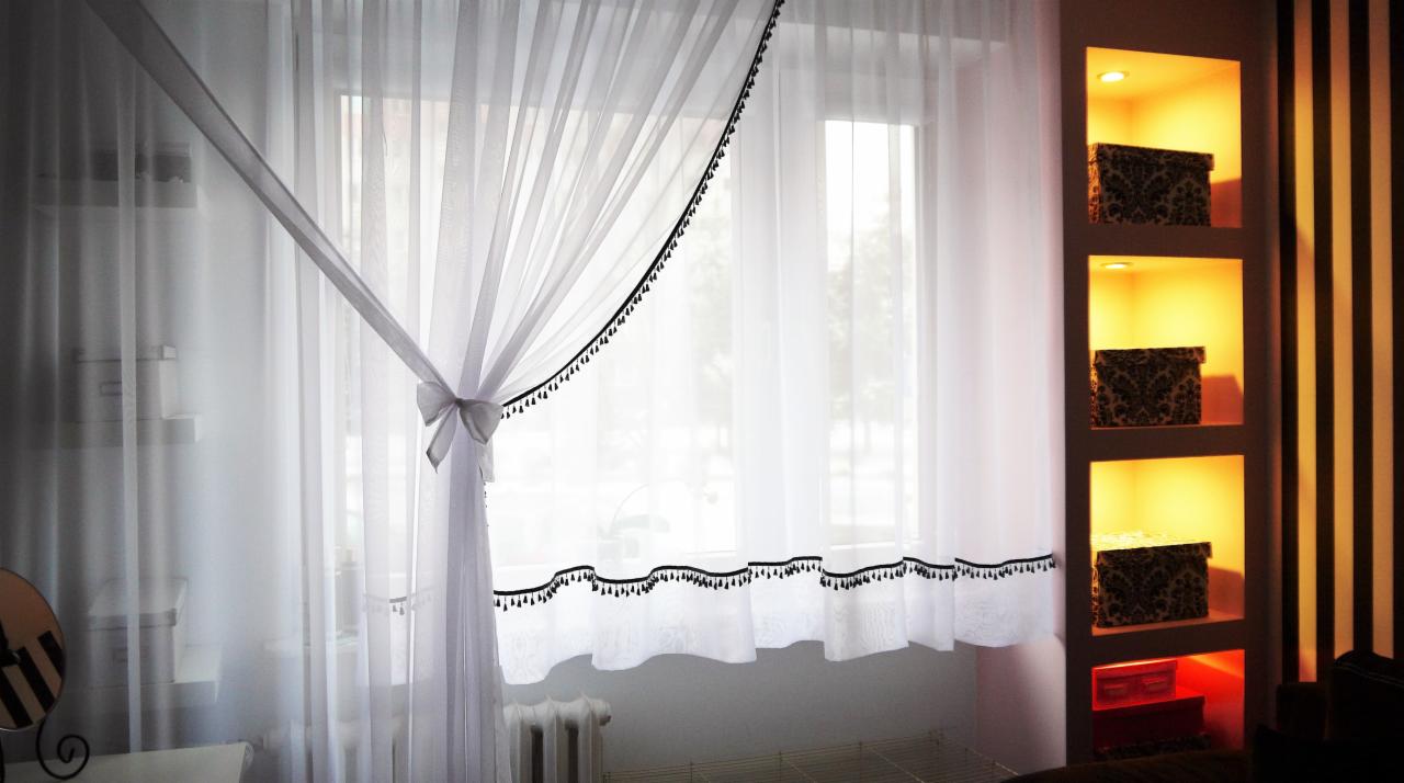 Delikatna, biała zasłona z czarnymi pomponami, upięta w kokardę, w jasnym wnętrzu z półkami i dekoracyjną ścianą w paski. Subtelny, elegancki design okna.