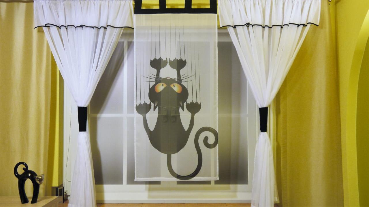 Okna z białymi, zwiewnymi zasłonami, po bokach upięte czarnymi pasami. Na środku roleta z nadrukiem czarnego kota drapiącego szybę. Obok stoi figurka kota.