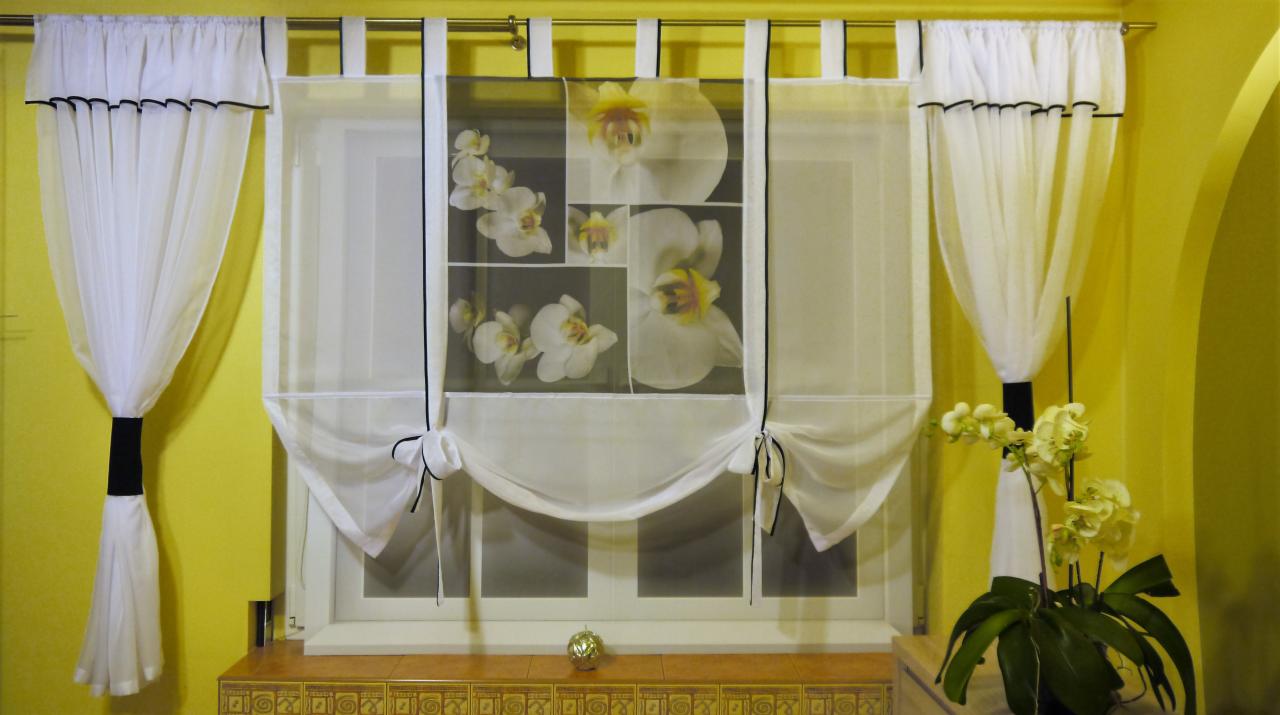Eleganckie, białe zasłony z czarnymi akcentami, w tym austriacka z motywem orchidei, dekorują okno na tle żółtej ściany, tworząc przytulną aranżację.