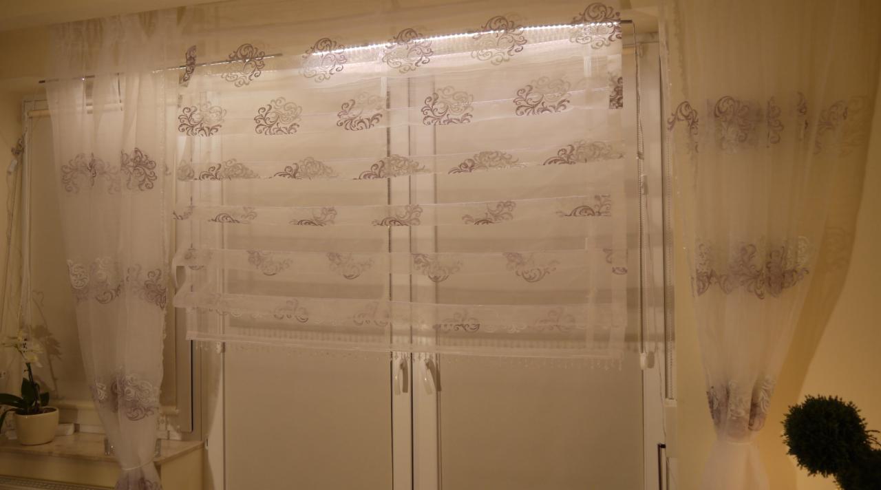 Delikatne, białe zasłony i roleta rzymska z subtelnym, srebrnym wzorem zdobią okno, tworząc elegancki wystrój wnętrza. Widoczne detale wykończenia.