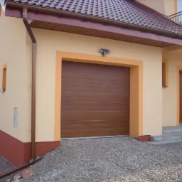 Zewnętrzny widok domu z brązową segmentową bramą garażową, otoczoną beżową ramą. Podjazd wysypany żwirem. Widoczna elewacja w odcieniach beżu i brązu.