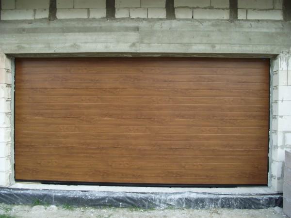 Zamknięta, brązowa brama garażowa segmentowa z poziomymi przetłoczeniami, w surowym otworze budynku z betonu komórkowego.