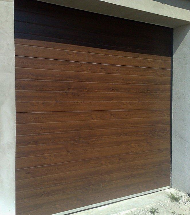 Zamknięta, brązowa brama garażowa segmentowa z poziomymi panelami imitującymi drewno, zamontowana w szarej, betonowej wnęce. Widoczne drobne elementy otoczenia.