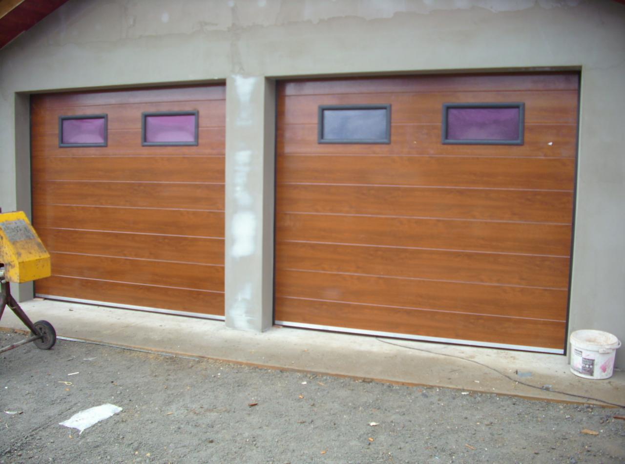 Dwie brązowe bramy garażowe segmentowe z poziomymi panelami i prostokątnymi okienkami, oddzielone betonowym słupkiem, na tle szarej ściany budynku.
