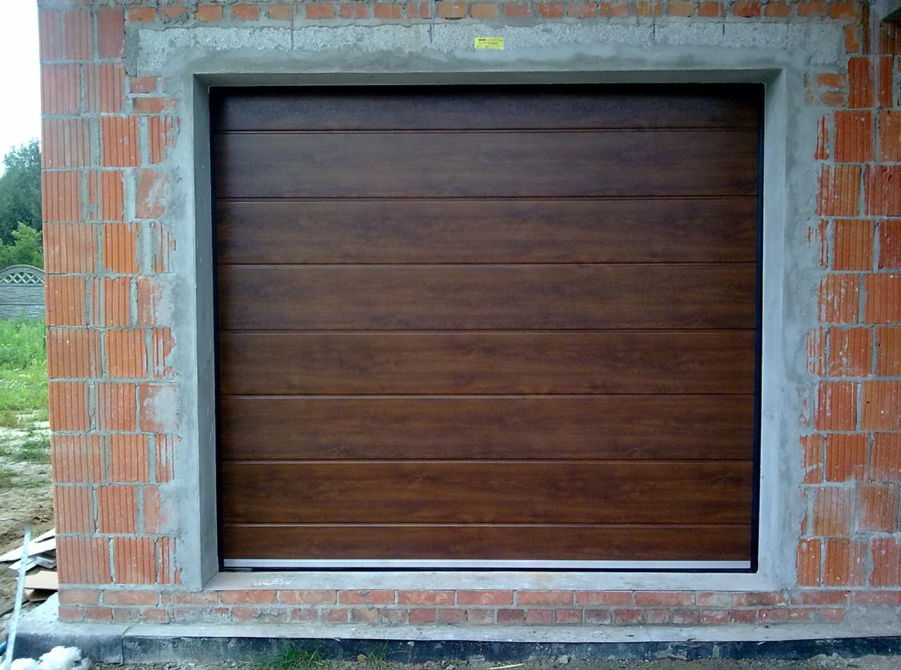 Segmentowa brama garażowa w kolorze drewna, widoczna w surowym otworze muru z cegły. Świeży montaż, brama zamknięta, widoczne detale konstrukcyjne.