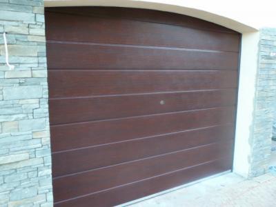 Ciemnobrązowa brama garażowa segmentowa, z poziomymi przetłoczeniami, zamontowana w jasnej elewacji z kamienia. Widok z boku.