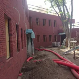 Ceglany budynek w trakcie murowania, widoczne otwory okienne z izolacją, czerwone rury na ziemi i sprzęt budowlany. Ujęcie z placu budowy.