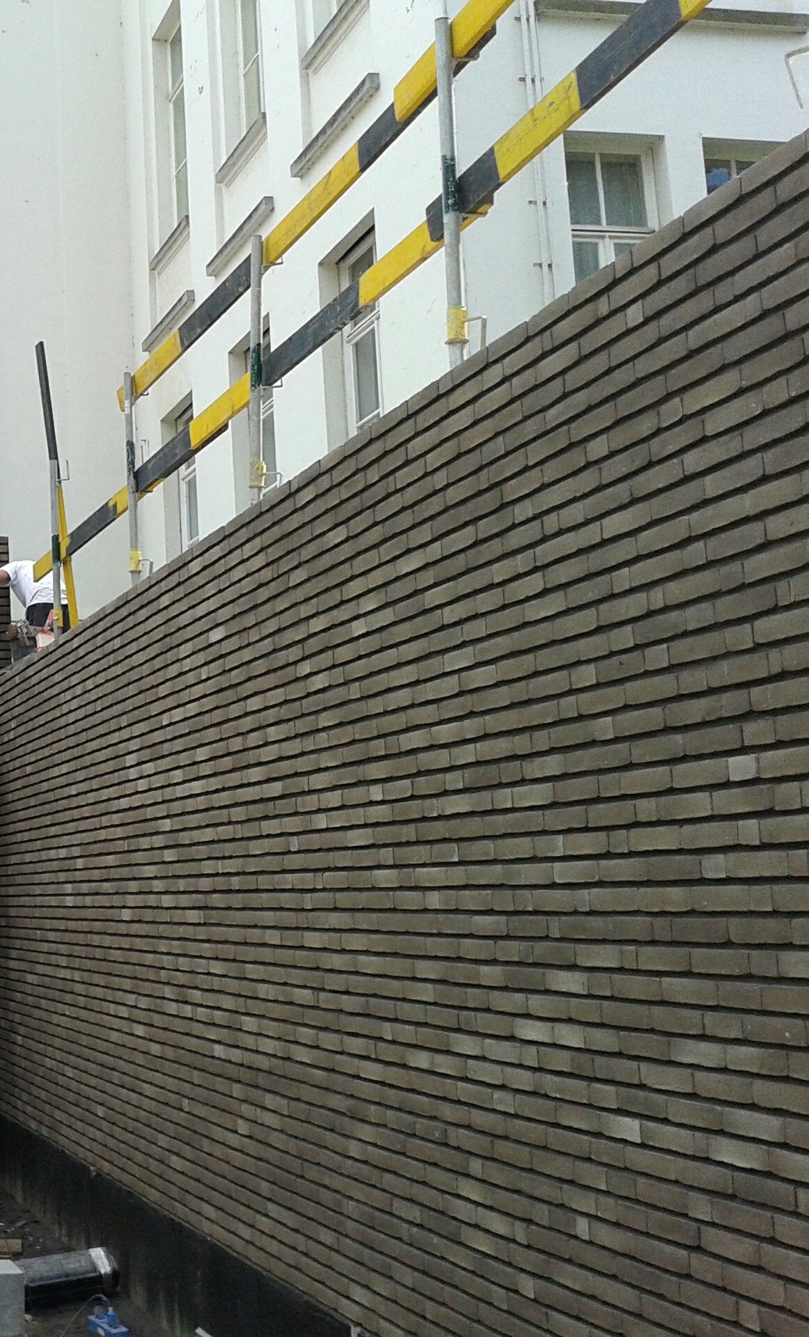 Nowo wymurowana ściana z cegły klinkierowej w odcieniach szarości, z widocznym rusztowaniem w tle. Cegły ułożone poziomo, tworzą regularny wzór. Detal ukończonej realizacji.