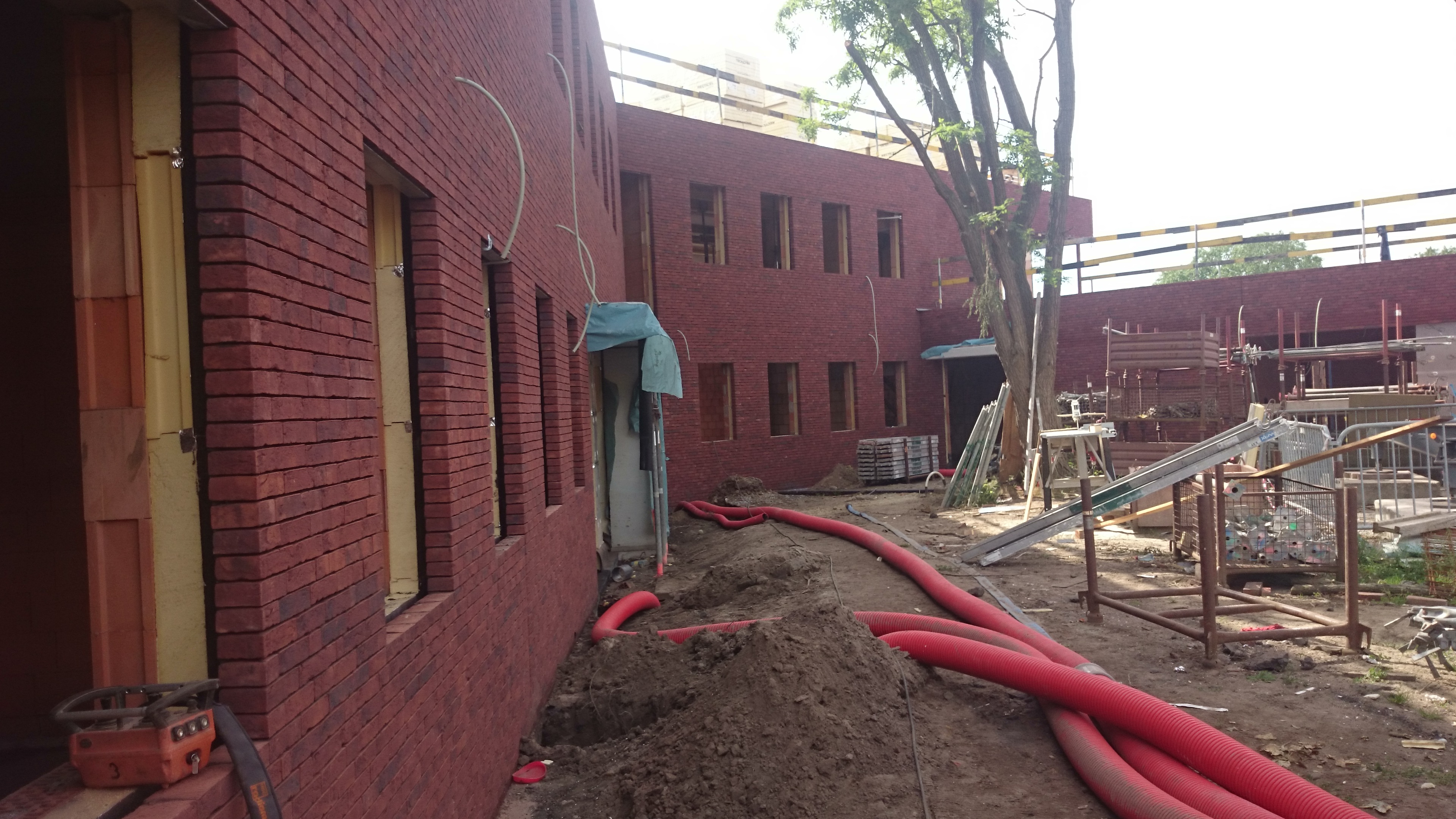 Ceglany budynek w trakcie murowania, widoczne otwory okienne z izolacją, czerwone rury na ziemi i sprzęt budowlany. Ujęcie z placu budowy.