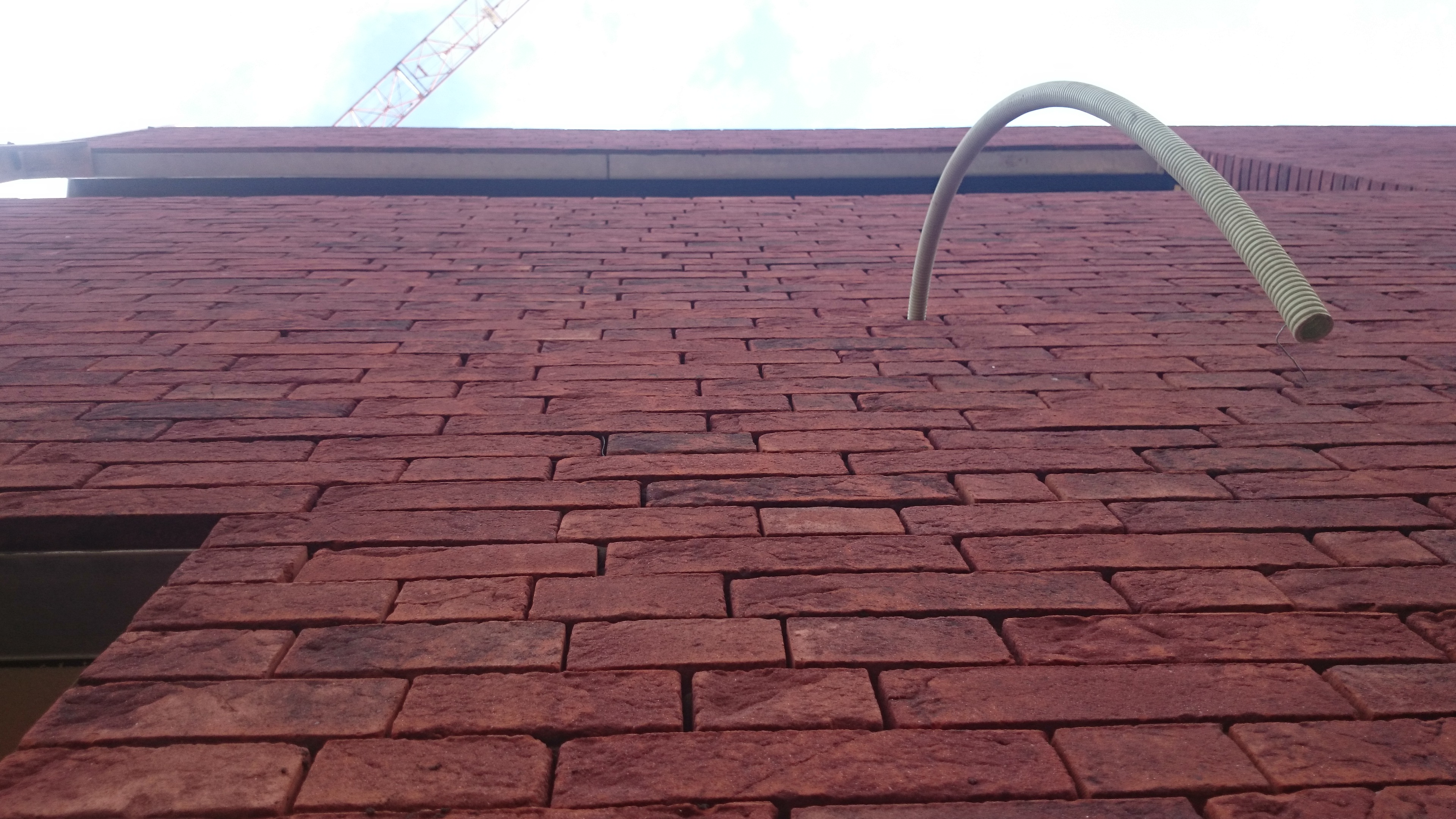 Ceglana ściana w Tychach z wystającą rurą instalacyjną, widok z dołu. Czerwona cegła, szara rura, budowa w toku, na górze widoczny dźwig.