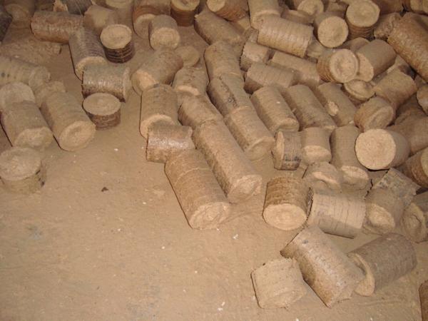 Stos cylindrycznych i okrągłych brykietów drzewnych w magazynie, widoczne ślady pyłu drzewnego na podłożu.