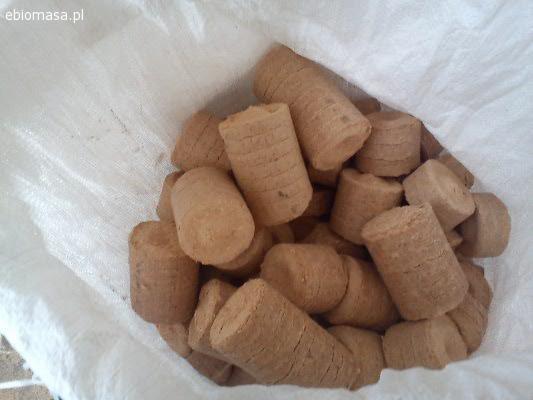 Jasnobrązowe brykiety w kształcie walców, ułożone w białym worku. Widoczne delikatne prążki na niektórych brykietach, sugerujące proces produkcji.