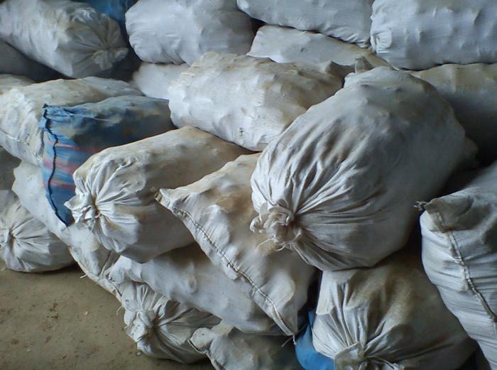 Stos worków z zawartością, prawdopodobnie trocin lub zrębków, ułożonych jeden na drugim. Niektóre worki są zabrudzone, a jeden ma niebieskie paski.