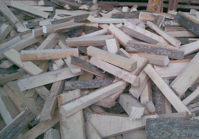 Stos pociętego drewna kominkowego z korą, ułożonego chaotycznie. Widoczne jasne drewno i fragmenty kory. W tle słabo widoczna drewniana konstrukcja.