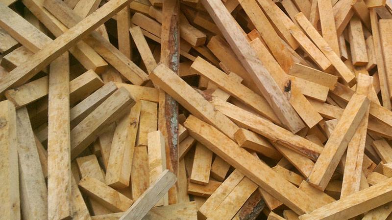 Stos pociętych kawałków drewna o jasnym odcieniu, ułożonych chaotycznie. Widoczne różne długości i grubości elementów, niektóre z korą. Surowe drewno tartaczne.