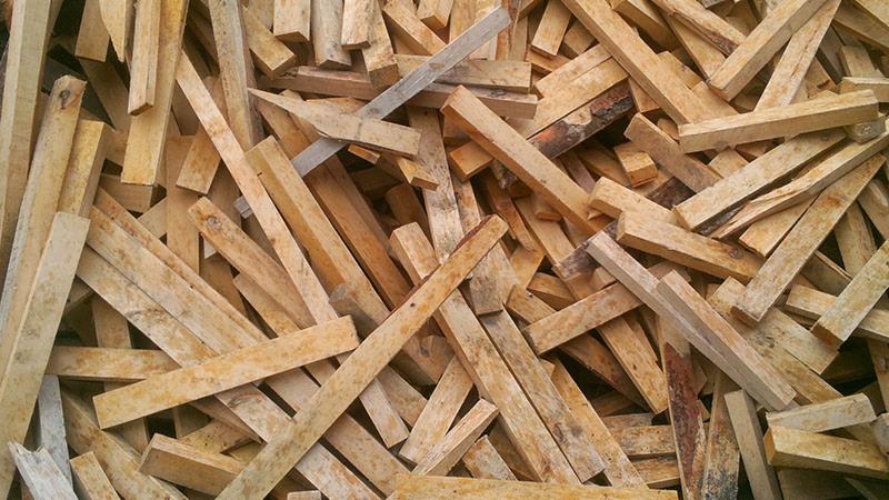 Stos pociętych listew z jasnego drewna ułożonych chaotycznie, widoczne sęki i naturalna faktura surowca. Nieregularne krawędzie i różna długość elementów.