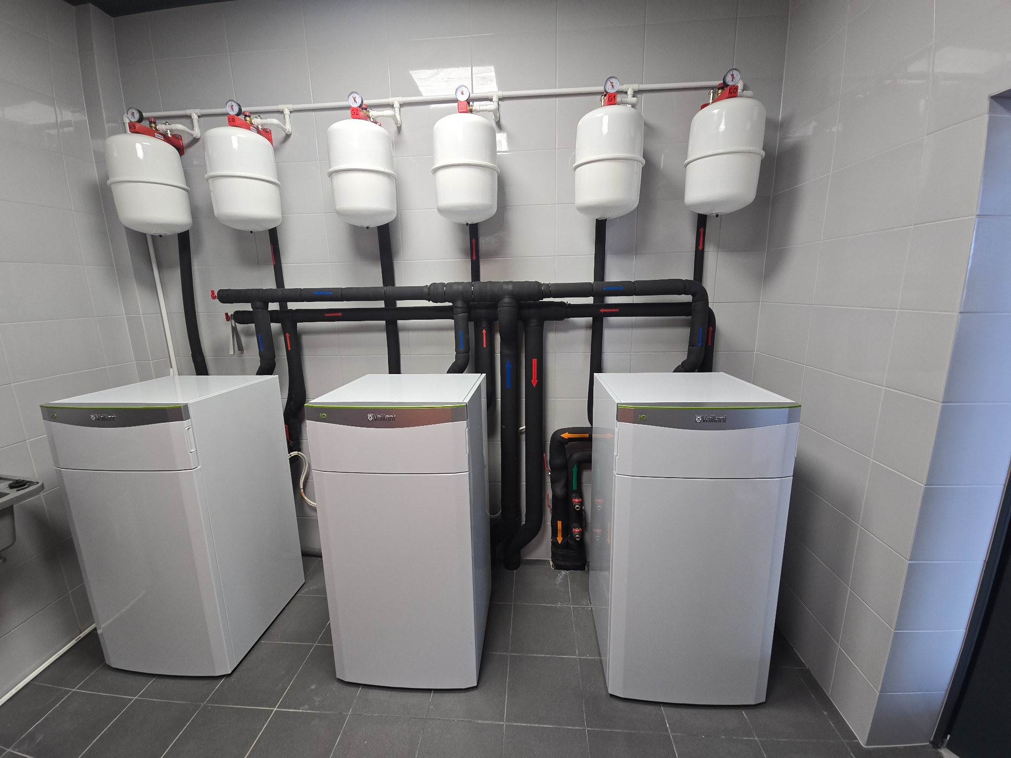 Trzy białe pompy ciepła Vaillant z czarnymi rurami i białymi zbiornikami wyrównawczymi w szarej, wykafelkowanej kotłowni. Widok z przodu, lekki kąt z lewej.