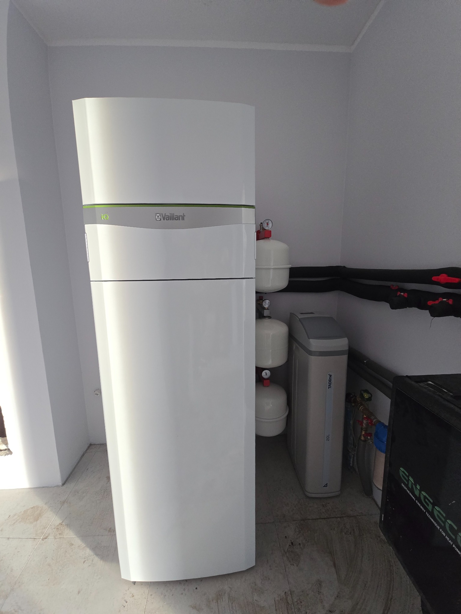 Biała pompa ciepła Vaillant w kotłowni, widoczne rury instalacyjne, zbiorniki wyrównawcze i zmiękczacz wody. Ujęcie z bliska, neutralne oświetlenie.