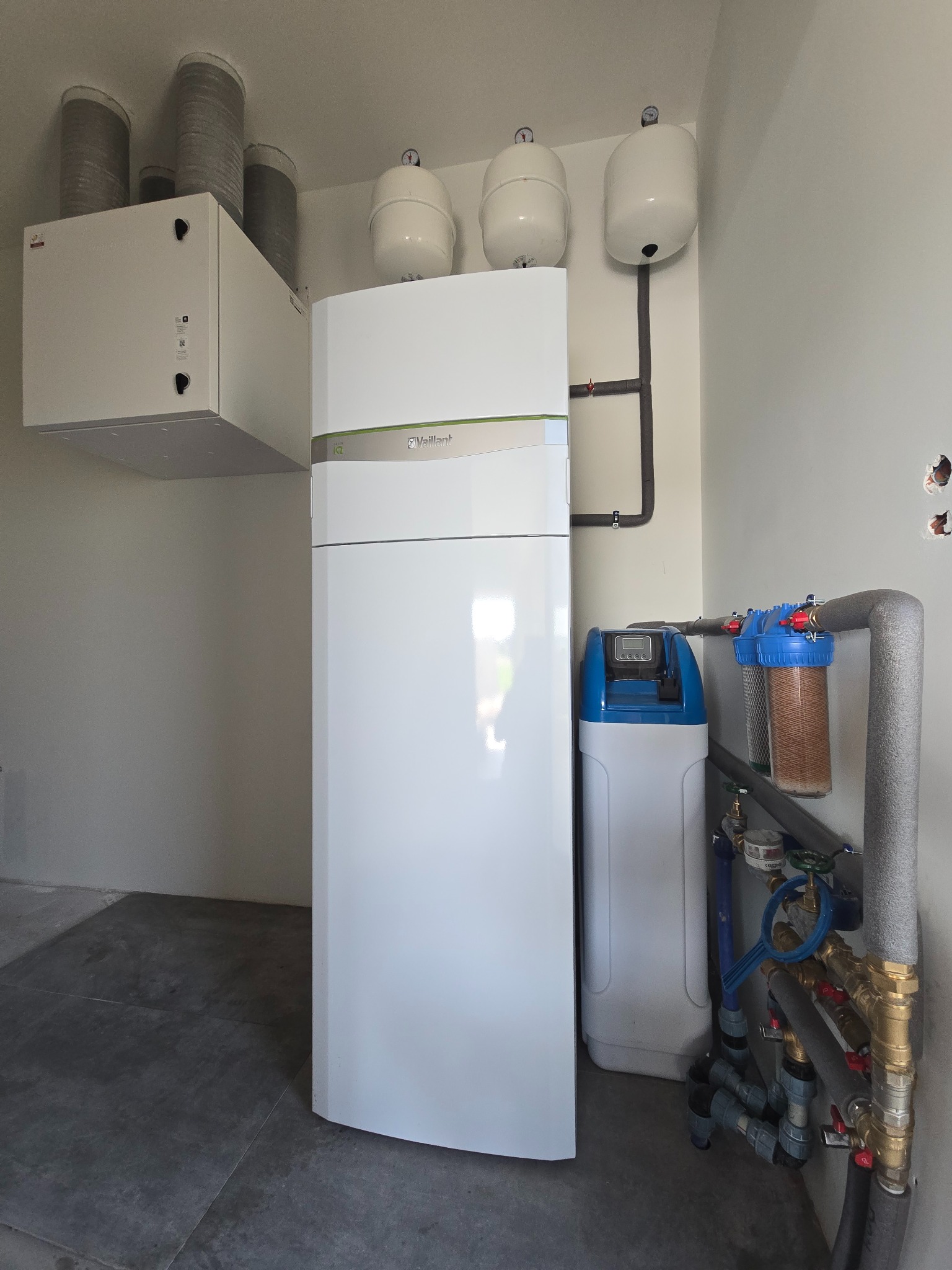Biała pompa ciepła Vaillant, zbiorniki wyrównawcze, filtry wody i zmiękczacz w kotłowni. Szare rury z izolacją. Widok na szarą podłogę i białe ściany.
