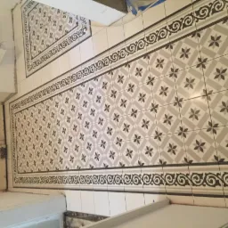 Ukończona podłoga w łazience wyłożona wzorzystymi, beżowo-szarymi płytkami z dekoracyjnym, ciemnym obramowaniem, widok z góry na fragment ściany i drzwi.