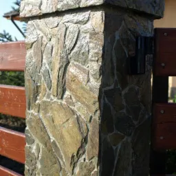 Kamienny słupek ogrodzeniowy z nieregularnych, brązowo-szarych płytek, połączony z drewnianym ogrodzeniem. Widoczna lampa. Słoneczny dzień.