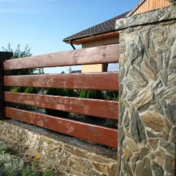 Drewniano-kamienne ogrodzenie posesji z brązowymi deskami i słupkami obłożonymi kamieniem w odcieniach szarości i brązu. Widok z zewnątrz, w tle zieleń.