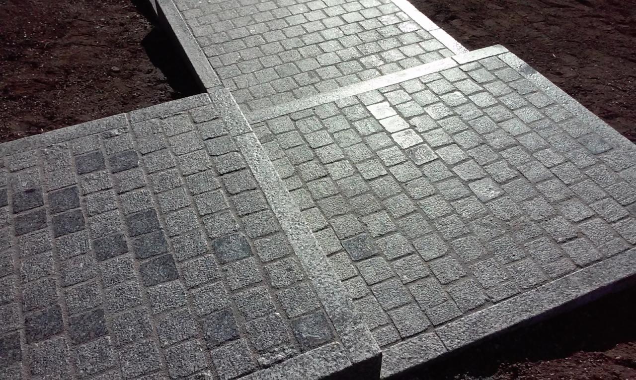 Ułożona kostka granitowa w odcieniach szarości, tworząca schody i chodnik. Widoczne regularne wzory kostki i obrzeża. Nierównomierne oświetlenie podkreśla fakturę kamienia.