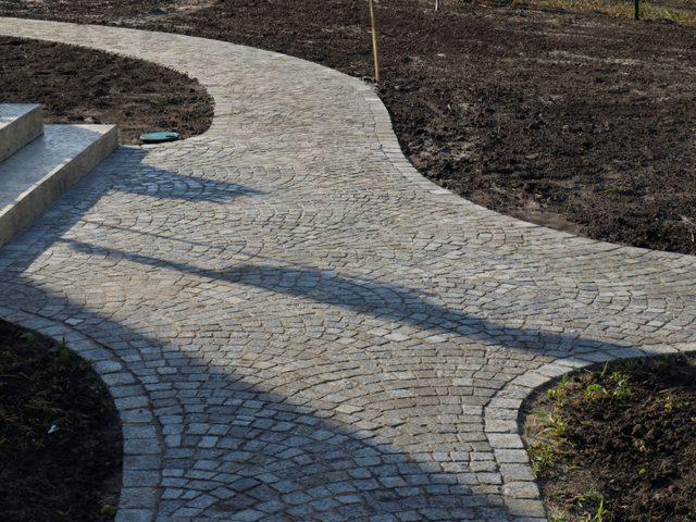 Granitowa ścieżka rozwidlająca się w ogrodzie, ułożona w łukowaty wzór. Widoczne schody z kostki i przygotowane rabaty ziemi.