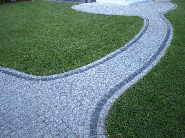 Granitowa ścieżka wijąca się przez trawnik, obrzeża z ciemnej kostki, jasna kostka ułożona w łukowaty wzór, widok z góry, równo skoszona trawa.