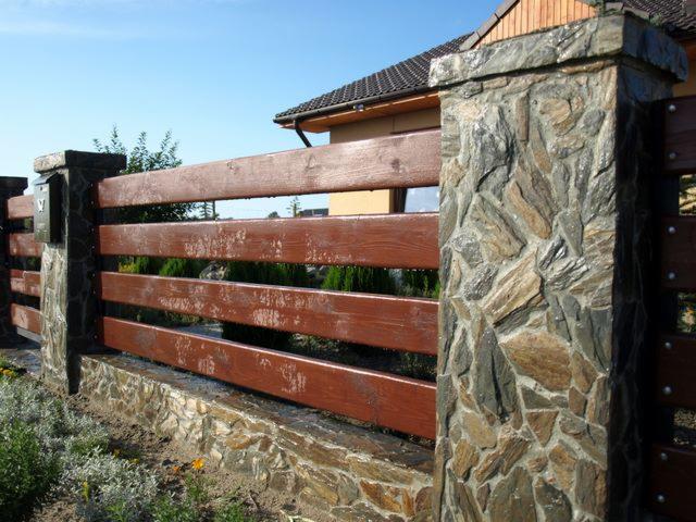 Drewniano-kamienne ogrodzenie posesji z brązowymi deskami i słupkami obłożonymi kamieniem w odcieniach szarości i brązu. Widok z zewnątrz, w tle zieleń.