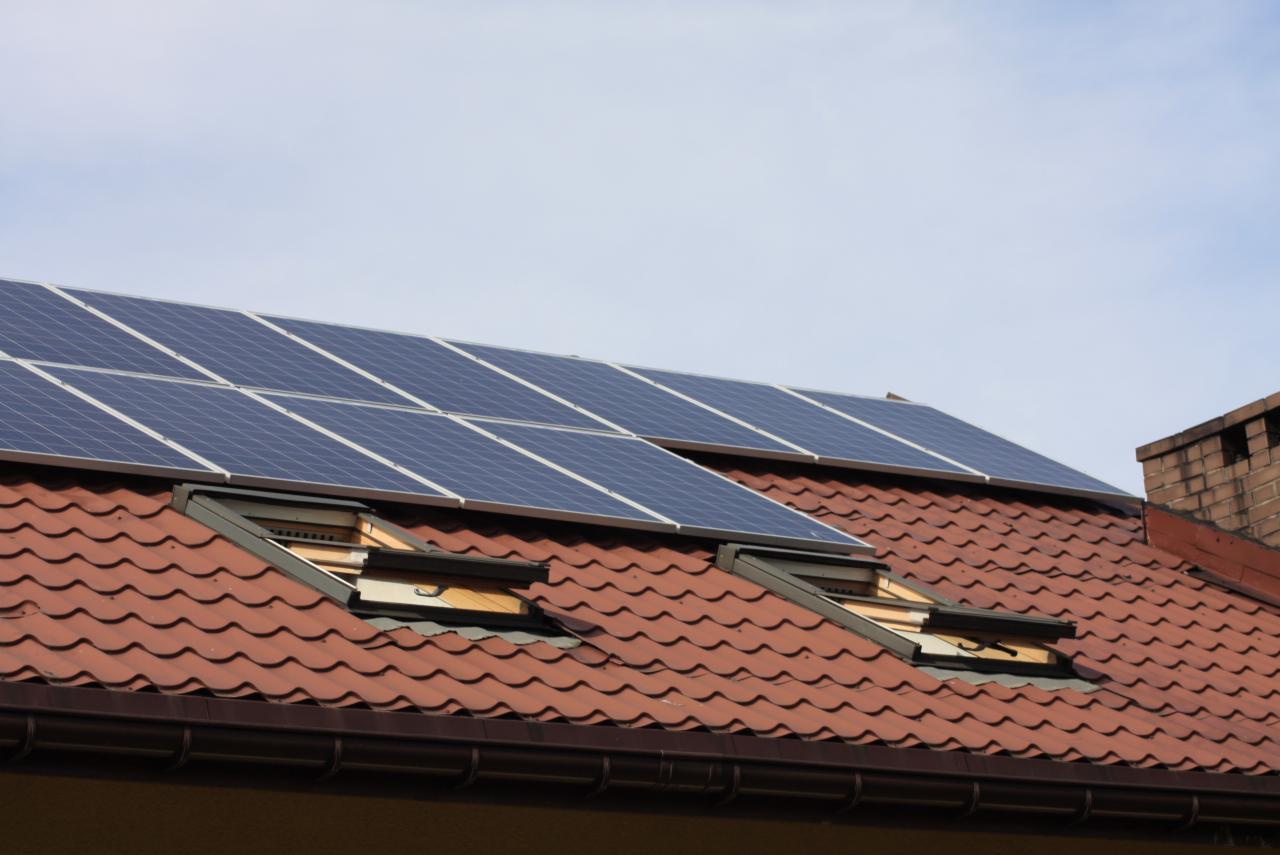 Czerwony dach z panelami słonecznymi i oknami dachowymi. Panele ułożone rzędami, widoczne detale konstrukcji. Ujęcie z dołu, błękitne niebo w tle.