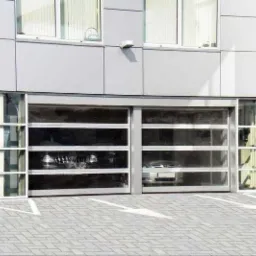 Szklana brama garażowa segmentowa z aluminiową ramą, częściowo otwarta, z widocznymi dwoma samochodami w środku, na tle szarej elewacji budynku.