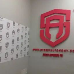 Czerwone logo firmy Street Autonomy w kształcie tarczy z wpisaną literą A, umieszczone na szarej ścianie nad adresem strony internetowej i nazwą firmy, z tapetą z powtarzającym się logo po lewej...